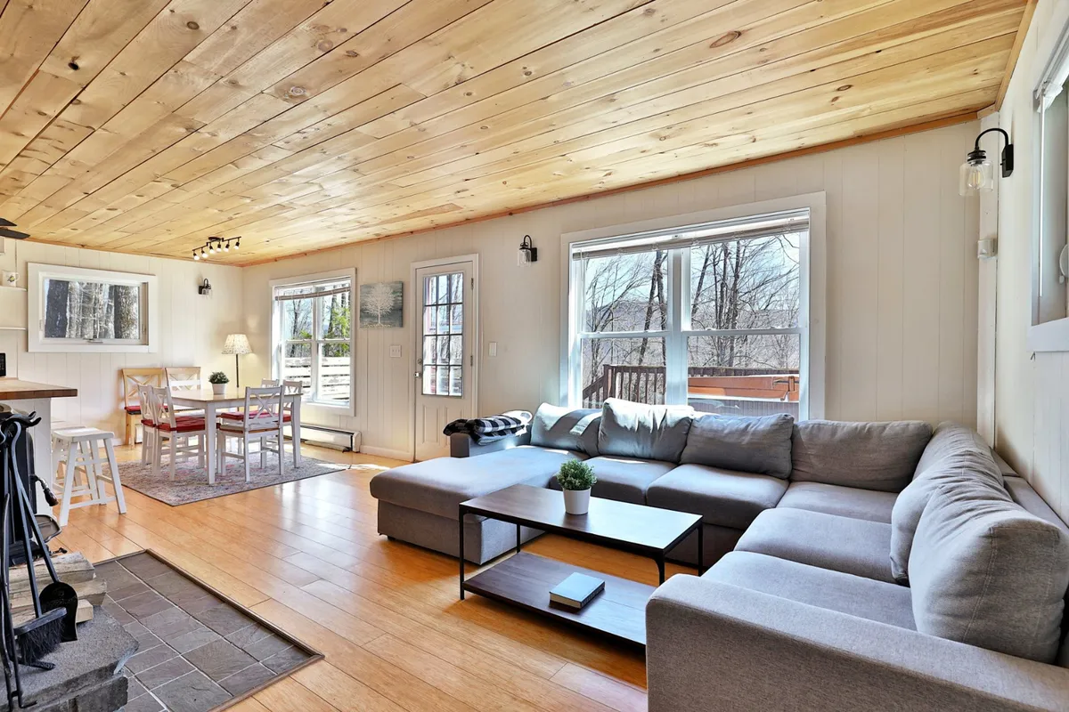 The image showcases a spacious, inviting living area with a large sectional sofa perfect for group gatherings, a family-style dining table, warm wooden ceilings, and ample windows framing serene natural views. The setting is filled with natural light, creating a cozy and welcoming ambiance ideal for relaxing or socializing after a day on the slopes.