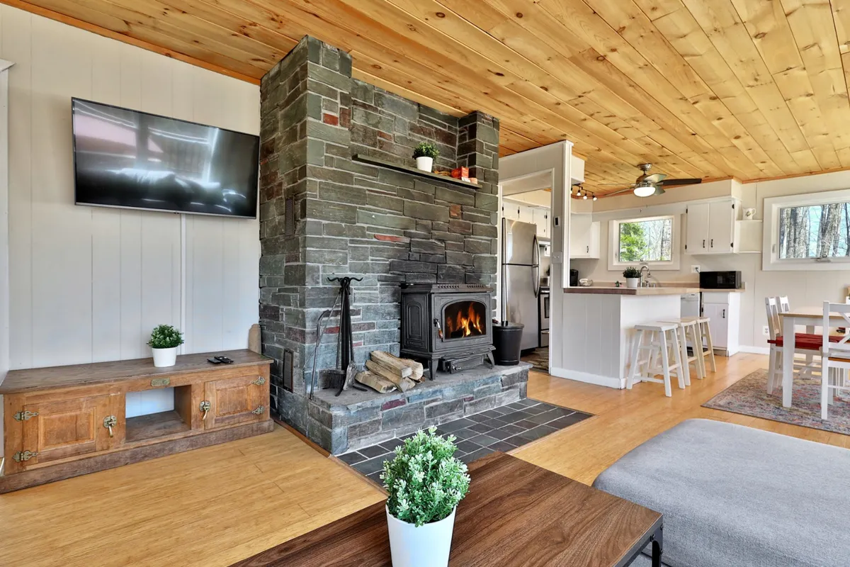 A wide view captures the living area with a stone fireplace, wooden ceiling, and a modern kitchen seamlessly connected to the dining table. The layout showcases the open-concept design, making it easy to see how the space encourages both cooking and gathering.