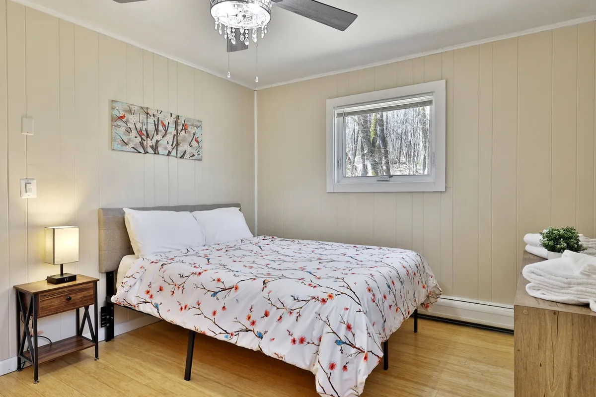 A serene bedroom with floral bedding on the bed, a nightstand with a lamp, and a window framing a view of the outdoors. Soft natural light fills the room, creating a peaceful atmosphere ideal for relaxation or a quiet moment with a book.