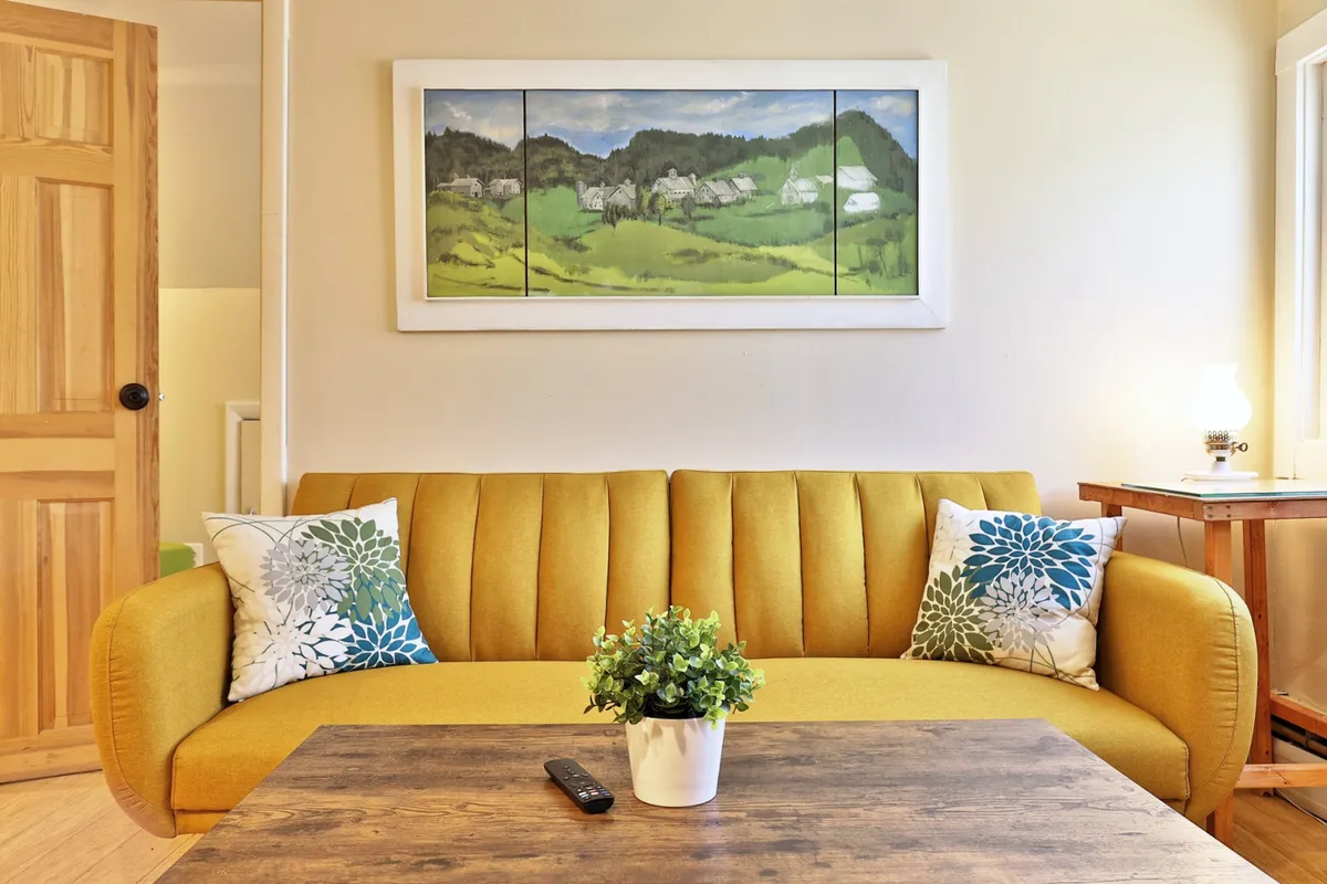 The image features a cheerful living area with a yellow couch, colorful decorative pillows, a wooden coffee table, lively artwork on the wall, and a small plant, creating a welcoming space ideal for social gatherings and group relaxation.