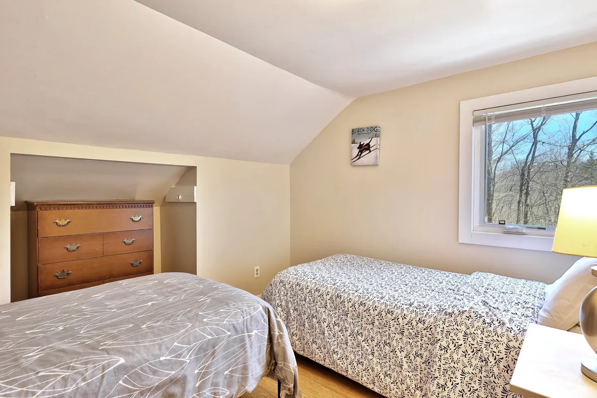 This image features a bedroom with two twin beds, a wooden dresser, and a window offering natural light. The room appears bright, tidy, and suitable for children or friends, with a calm and inviting atmosphere.