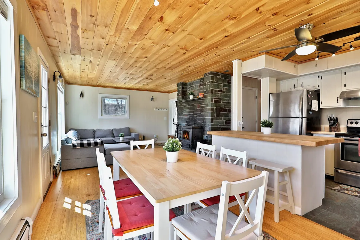 A cozy, inviting living room with a wooden dining table, a comfortable sofa set near a rustic stone fireplace, and expansive windows that let in natural light. The layout is perfect for family dinners, game nights, or relaxing by the fire after a day of exploring.