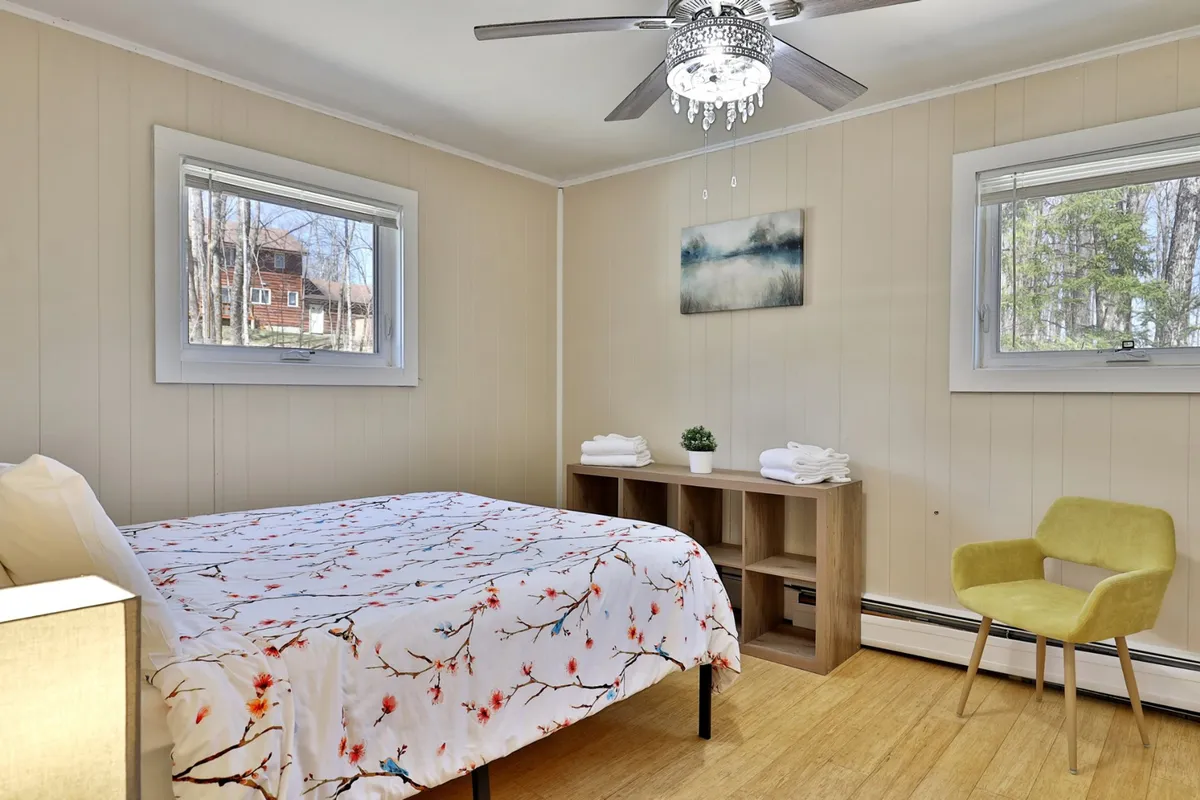 A calm and inviting bedroom with floral bedding, a wooden shelf, green plant, yellow chair, and wall art. The natural light filtering in sets a peaceful tone for moments of rest and quiet reflection.