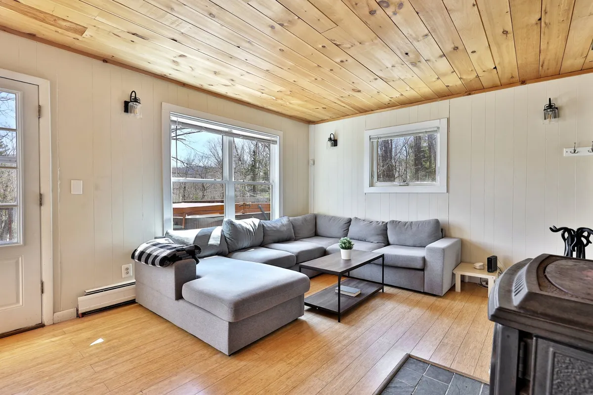 A spacious living area with a sectional sofa, coffee table, large windows letting in ample sunlight, and a wooden ceiling. The setting is comfortable and serene, inviting relaxation, reading, and social gatherings.