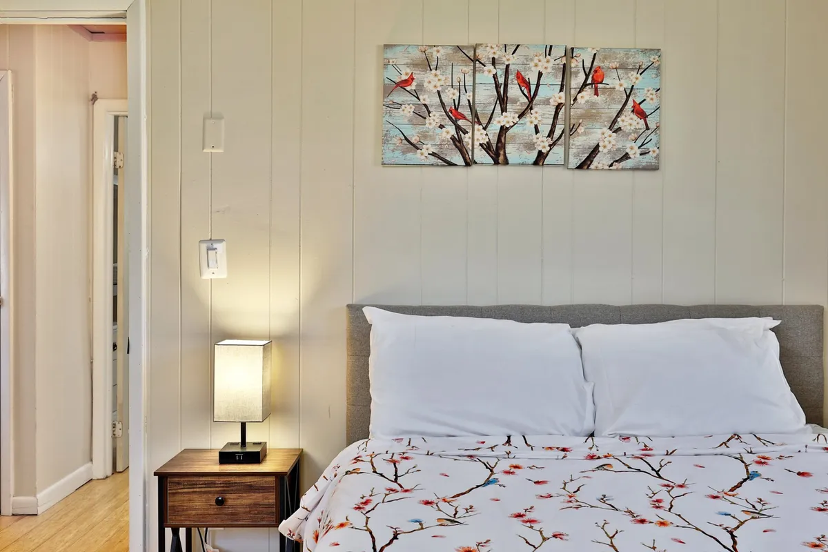 A tranquil queen bedroom with tasteful artwork above the bed, a nightstand with a lamp, and soft natural lighting filtering in. The mood is restful and serene, ideal for relaxation and quiet moments.
