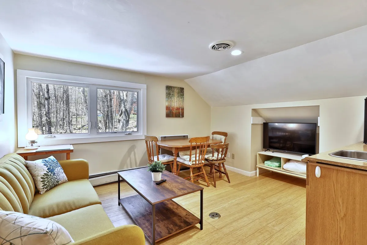 A spacious, light-filled living area featuring a green sofa, a dining table with chairs, a coffee table adorned with a plant, and a large window that offers a view of the surrounding trees. The setting suggests a warm, communal space perfect for family relaxation and socializing while keeping an eye on the outdoors.