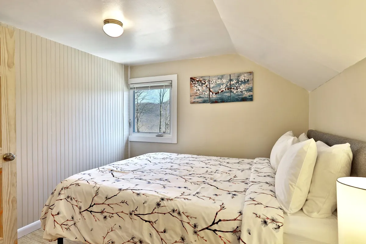 A tranquil bedroom with a floral bedspread, a window overlooking the outdoors, and nature-inspired artwork on the wall. The setup suggests a peaceful retreat where guests can unwind or work in quiet comfort, with easy access to nature just outside.