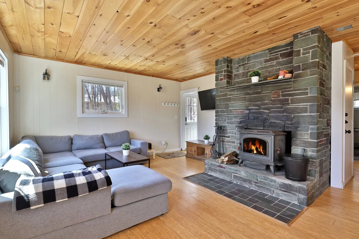 This image features the main living room with a prominent stone fireplace, a comfortable sectional sofa, and a warm wooden ceiling. The scene is set for relaxation and socializing, enhanced by natural light streaming through the windows. The fireplace is stocked with wood, and the atmosphere is inviting, perfect for gathering after a day outdoors.